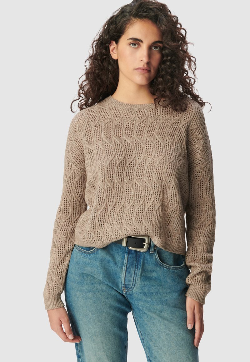 Frau mit lockigem Haar, die einen beigen, strukturierten Pullover und blaue Jeans mit einem schwarzen Gürtel trägt, steht vor einem schlichten Hintergrund.