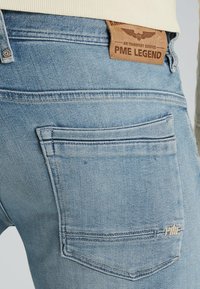 Light blue denim jeans feature a textured finish, a leather patch with "PME LEGEND," and lighter stitching. Back pocket design is minimal.