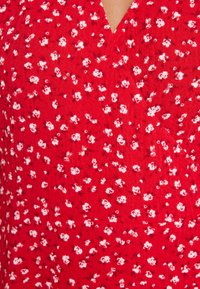Červená látka s květinovým vzorem obsahující malé bílé květy. Textura vypadá měkce, má design s výstřihem do V a mírně zmačkaný vzhled.