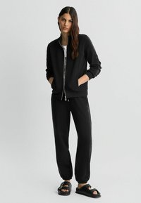 Veste bomber noire avec fermeture éclair, tissu texturé, poignets côtelés ; associée à un pantalon de jogging noir avec ourlets élastiques et accents sur les côtés.