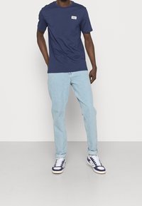 Camiseta de algodón azul marino con un logo blanco de FILA en el pecho, combinada con jeans azul claro y zapatillas navy con acentos blancos.