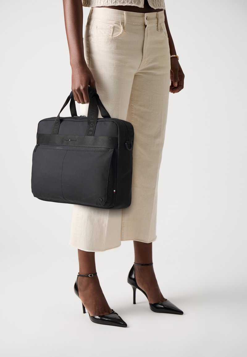 Personne portant un pantalon large crème et des talons noirs tient à côté d'elle une mallette carrée noire avec des poignées.