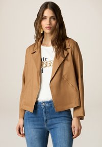 Bruine, cropped jas met een geknipte kraag en open voorkant. Gemaakt over een witte shirt met gouden pailletten tekst. Blauwe denim jeans maken de look compleet.