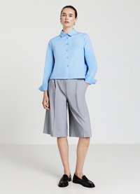 Hellblaues Hemd mit Knopfleiste, langen Ärmeln und Brusttasche, kombiniert mit grauen Falten-Shorts; schwarze Loafers vollenden den Look.