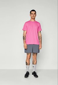 Rosa kurzärmliges Sportshirt mit grauen Shorts, trägt schwarze Sneakers und graue Kniestrümpfe. Hat Tattoos an den Armen. Einfache Hintergrund.