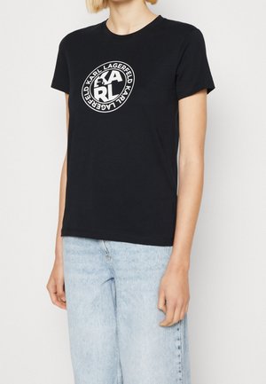 Persoon die een zwart T-shirt met korte mouwen draagt met een wit rond Karl Lagerfeld-logo op de borst en lichtblauwe spijkerbroek.