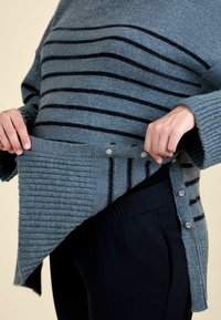 Kék csíkos pulóver bordázott textúrával, ezüst gombokkal és réteges kialakítással. Sötét nadrággal párosítva a kontraszt érdekében.