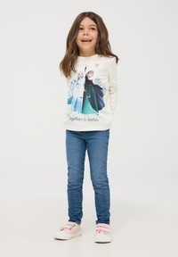 Bluzka z długim rękawem w kolorze kremowym z grafiką dwóch postaci w kolorowych sukienkach; połączona z niebieskimi dżinsami i pastelowymi sneakersami, na jednolitym tle.