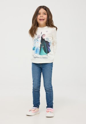 Maglietta a maniche lunghe color crema con una grafica di due personaggi in abiti colorati; abbinata a jeans blu e sneakers pastello, su uno sfondo semplice.