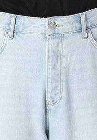 Light blue denim jeans feature a five-pocket design, a metal button closure, and tan stitching along the seams. Smooth texture and straight fit.