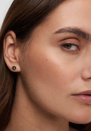 Nahaufnahme eines Ohrs einer Frau mit einem goldenen Ohrstecker mit grünem Edelstein, sichtbar sind ein Teil ihres Auges, ihrer Augenbraue und ihrer Lippen.