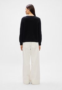 Juodas tekstūruotas megztinis su apvalia iškirpte, derinamas su baltomis plačiomis kelnėmis, turinčiomis du užpakalinius kišenes ir matomą rankogalių apdailą apačioje.