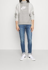 Šedá mikina s kapucí s bílým logem Nike a šňůrkami, v kombinaci s modrými slim-fit džínami a bílými teniskami s černými doplňky.