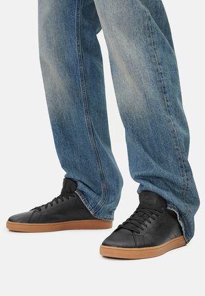 Persona, kas valkā vaļīgas zilas džinsas un melnus ādas sporta apavus ar brūnām gumijas zolēm, stāv uz balta fona.