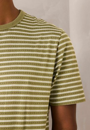 Felső test rész olívazöld rövid ujjú pólóban, fehér és fekete vízszintes csíkokkal és apró pöttyös mintával, semleges háttéren.