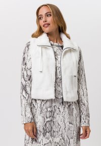 Witte faux fur cropped gilet met een rits aan de voorkant en trekkoorden, gedragen over een lange mouwen jurk met slangenprint in grijze en witte tinten.