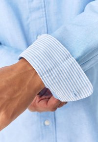 Lichtblauw overhemd met een button-down kraag en een contrasterende wit-lichtblauw gestreepte manchet. De stof lijkt glad met een subtiele textuur.