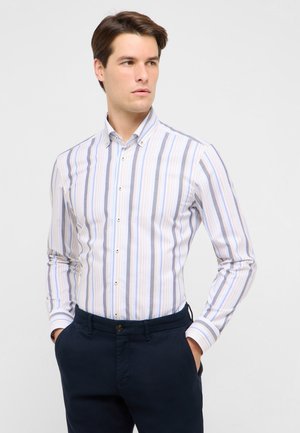 Ung mand iført en lyseblå, hvid og beige stribet skjorte med knapper og mørkeblå bukser, står med hænderne i lommerne og kigger til siden.