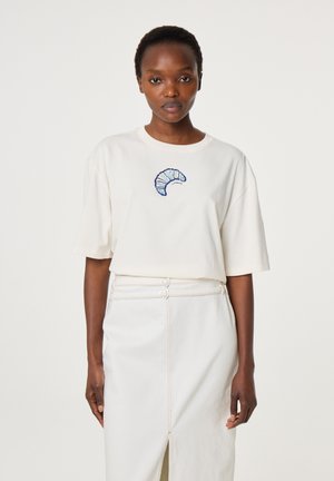 Vrouw draagt een wit t-shirt met een blauwe croissantprint en een witte rok met een split aan de voorkant en een dubbele hartvormige riemgesp.