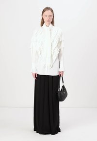 Blusa blanca con volantes y cuello atado, combinada con una falda larga negra y fluida. La modelo sostiene un bolso negro pequeño con correa para el hombro.