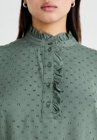 Kobieta ubrana w zieloną bluzkę o teksturowanym wykończeniu z guzikami i falbanką przy kołnierzyku oraz wzdłuż przodu.