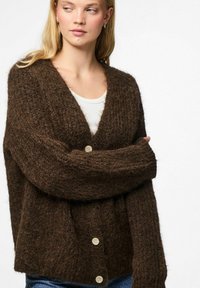 Blond naine pruunis karvases nööbitud kardiganis valge särke kandes, käed risti ja vaadates külje poole ühtlase tausta ees.