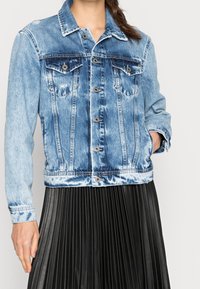 Denim jacket in light blue, featuring a classic design, two chest pockets, and silver button closures. Worn over a pleated black skirt.