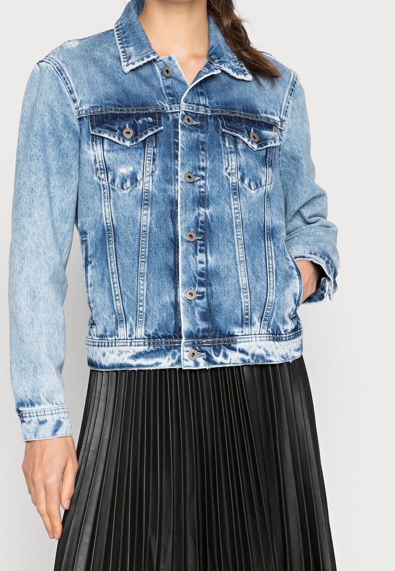 Denim jacket in light blue, featuring a classic design, two chest pockets, and silver button closures. Worn over a pleated black skirt.