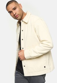 Beige Cord-Jacke mit Hemdblende, Druckknöpfen und subtilen horizontalen Streifen. Getragen über einem schwarzen Hemd und kombiniert mit grauen Hosen.