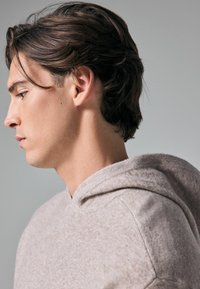 Hellgrau, strukturiertes Kapuzenshirt aus weichem Stoff mit einer vorderen Tasche. Verfügt über eine Kapuze mit Kordelzug und einen lässigen Schnitt. Auf einem Modell im Profil gezeigt.