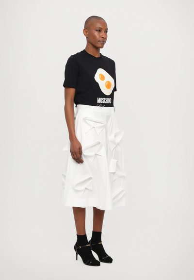 T-shirt negru cu grafică de ou alb și text "Moschino", asortat cu o fustă albă cu detalii din material alb protruzente. Sandale cu toc înalt negre.