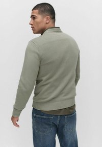 Ljusgrön stickad tröja med ribbat avslut och muddar, med en något upphöjd krage, bärs över en mörk skjorta och blå jeans.