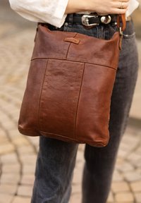 Hnedá kožená crossbody taška s hladkou textúrou, obdĺžnikovým tvarom a jemnými detailmi šitia. Prispôsobiteľný popruh pripevnený na vrchu.