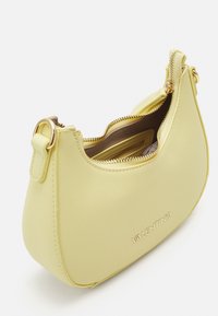 Žlutá crossbody taška vyrobená z texturovaného syntetického materiálu. Má zaoblený tvar, zlaté kování a zipové zapínání. Název značky je embosovaný.