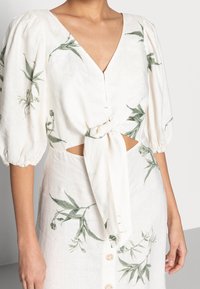 Blouse en lin blanc imprimée de motifs botaniques verts, manches bouffantes, encolure en V et un détail nœud à l'avant. Présente des découpes à la taille et des boutons.