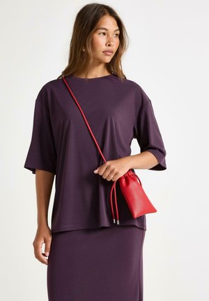 Fialový oversized top s krátkými rukávy, zkombinovaný s přiléhavou fialovou sukní. Doplňky zahrnují malou červenou tašku na stažení s hladkým povrchem.