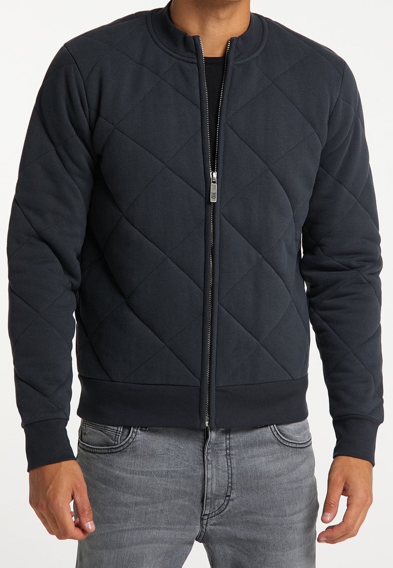 Vadderad bomberjacka i mörk marinblå, med rundad krage, dragkedja fram, ribbade muddar och midja, med en slät yta.