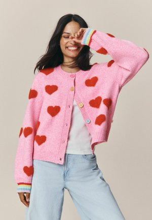 Young woman smiling with eyes closed wears a pink cardigan with red hearts and multicolor cuffs, white top, and light blue jeans.