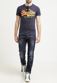 Námořnická modrá tričko s oranžovým a žlutým grafickým textem, v kombinaci s tmavými džínami a černobílými teniskami, v chůzi.
