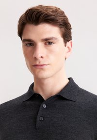 Dunkelgraues Poloshirt aus weichem Stoff, mit einem kragenähnlichen Ausschnitt und zwei dunklen Knöpfen. Nahaufnahme der Schultern und des Gesichts des Modells.