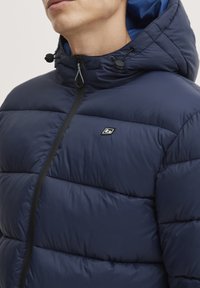 Námořnický modrý zateplený kabát s předním zipem, elastickou kapucí a nastavitelnými šňůrkami. Má vodorovné prošívání a malé logo.