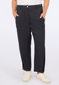 Osoba w czarnych spodniac h w prążki z bocznymi kieszeniami i białych sneakersach, stojąca z rękami w kieszeniach.