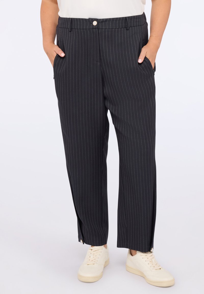 Osoba w czarnych spodniac h w prążki z bocznymi kieszeniami i białych sneakersach, stojąca z rękami w kieszeniach.