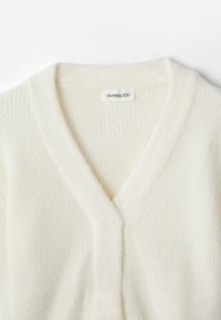V-hals cardigan i blødt cremefarvet materiale, med en ribbet tekstur og minimal mærkeetiket. Enkel, alsidig design.