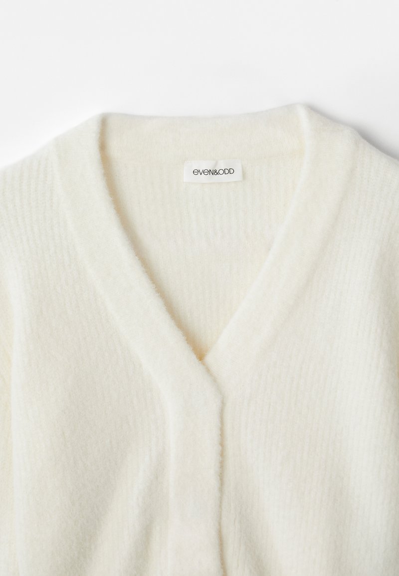 V-hals cardigan i blødt cremefarvet materiale, med en ribbet tekstur og minimal mærkeetiket. Enkel, alsidig design.