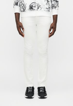 Moški v belih ozkih hlačah in črnih supergah z belimi vezalkami stoji z rokami sproščenimi ob telesu na belem ozadju.