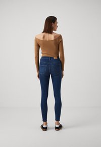 Top castanho sem mangas com textura de canelado, combinado com jeans justos azul-escuro, apresentando bolsos traseiros e um rótulo de couro bege na cintura.