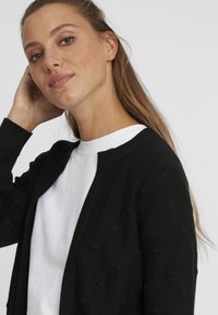 Cardigan noir avec des points texturés, à l'avant ouvert, avec des poignets et un col côtelés. Porté par-dessus une chemise blanche, affichant un design simple et moderne.