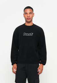 Sort sweatshirt lavet af blødt stof, med rund halsudskæring og et centralt "huf." logo i hvid. Båret med sorte cargobukser.