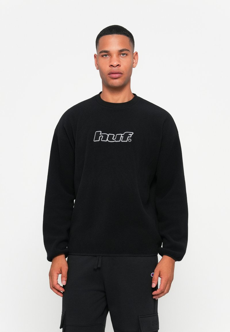 Sudadera negra hecha de una tela suave, con cuello redondo y un logo central "huf." en blanco. Combinada con pantalones cargo negros.